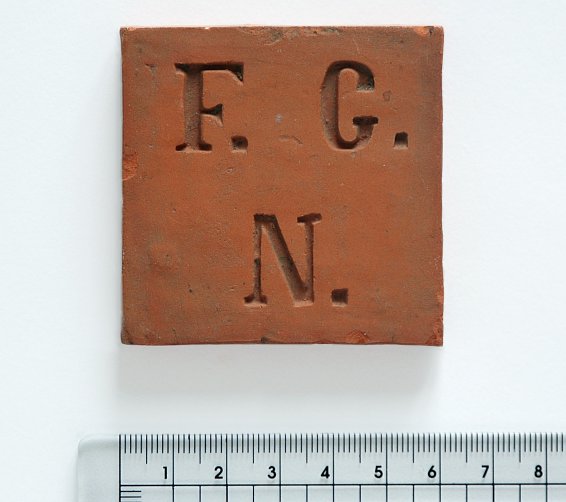 Eine quadratische Tontafel mit den Buchstaben F, G und N, darunter ein Lineal, die Kantenlänge beträgt 6 cm